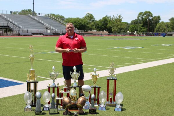 Chad stands on a football field holding his LJD Community Leader Award Trophy while his football trophies stand on the ground around him.